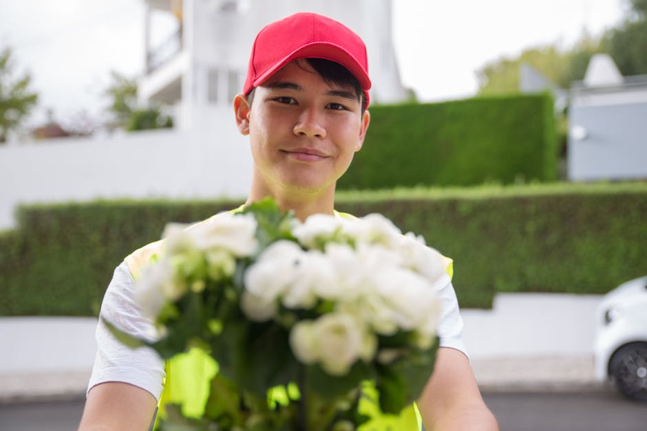 A smiling young delivery person wearing a red cap and a neon yellow vest is standing outdoors in a residential area, holding out a bouquet of fresh white flowers with green foliage. The bouquet is wra