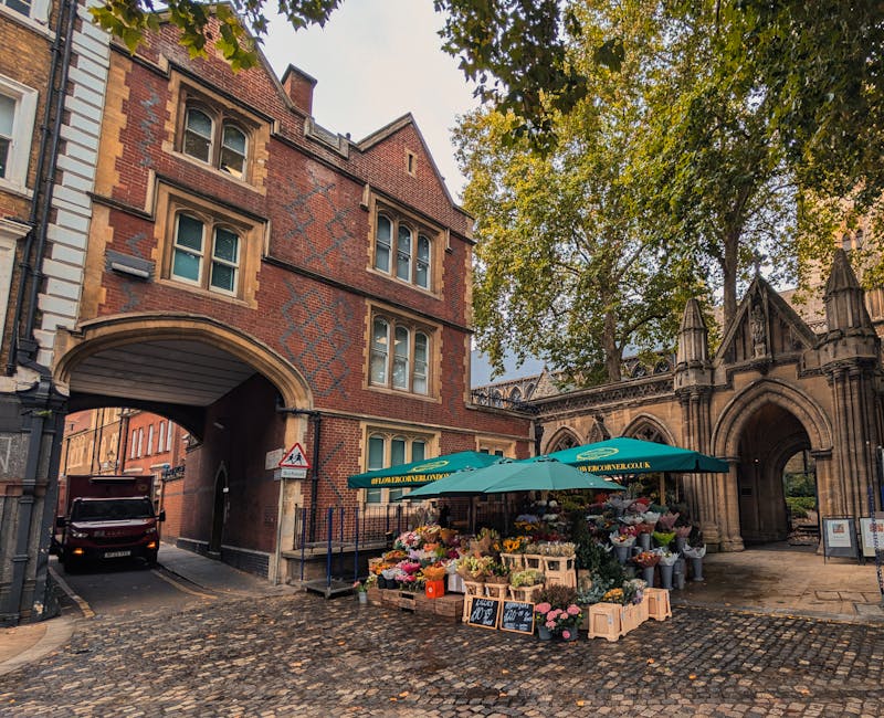 A vibrant outdoor flower stall situated on a cobblestone street near an archway and historic brick buildings. The stall is shaded by large green umbrellas and displays a wide variety of fresh flowers 