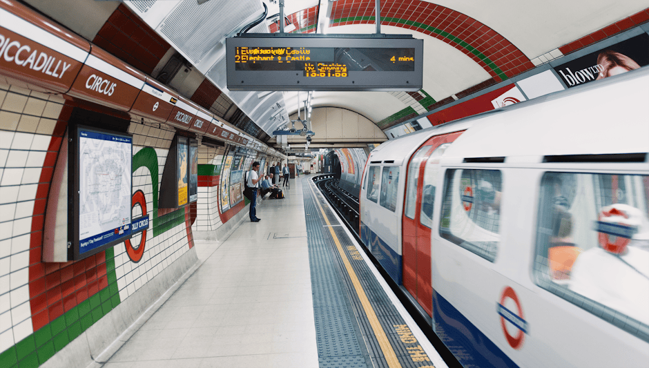 A London Underground station platform with tiled walls painted in white, red, and green, featuring digital departure boards overhead displaying upcoming train information. A train adorned with the ico