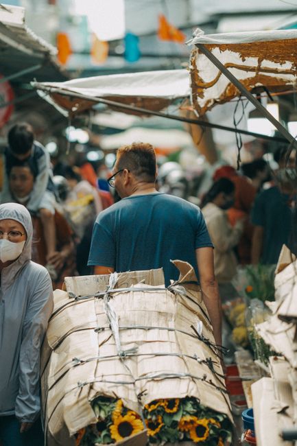 A busy outdoor flower market scene with a man wearing a blue shirt carrying a large bundle of fresh sunflowers wrapped in paper. Surrounding him are various vendors and shoppers, some wearing face mas