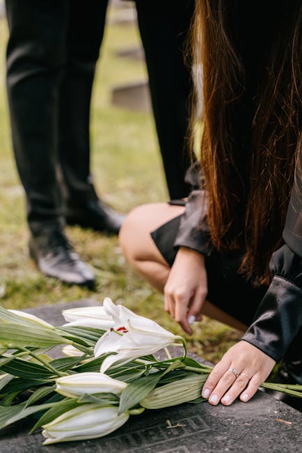 A person dressed in black kneels at a graveside, arranging a floral tribute of white lilies with long green stems and leaves, resting on a dark stone surface. The lilies are fresh, with fully opened p